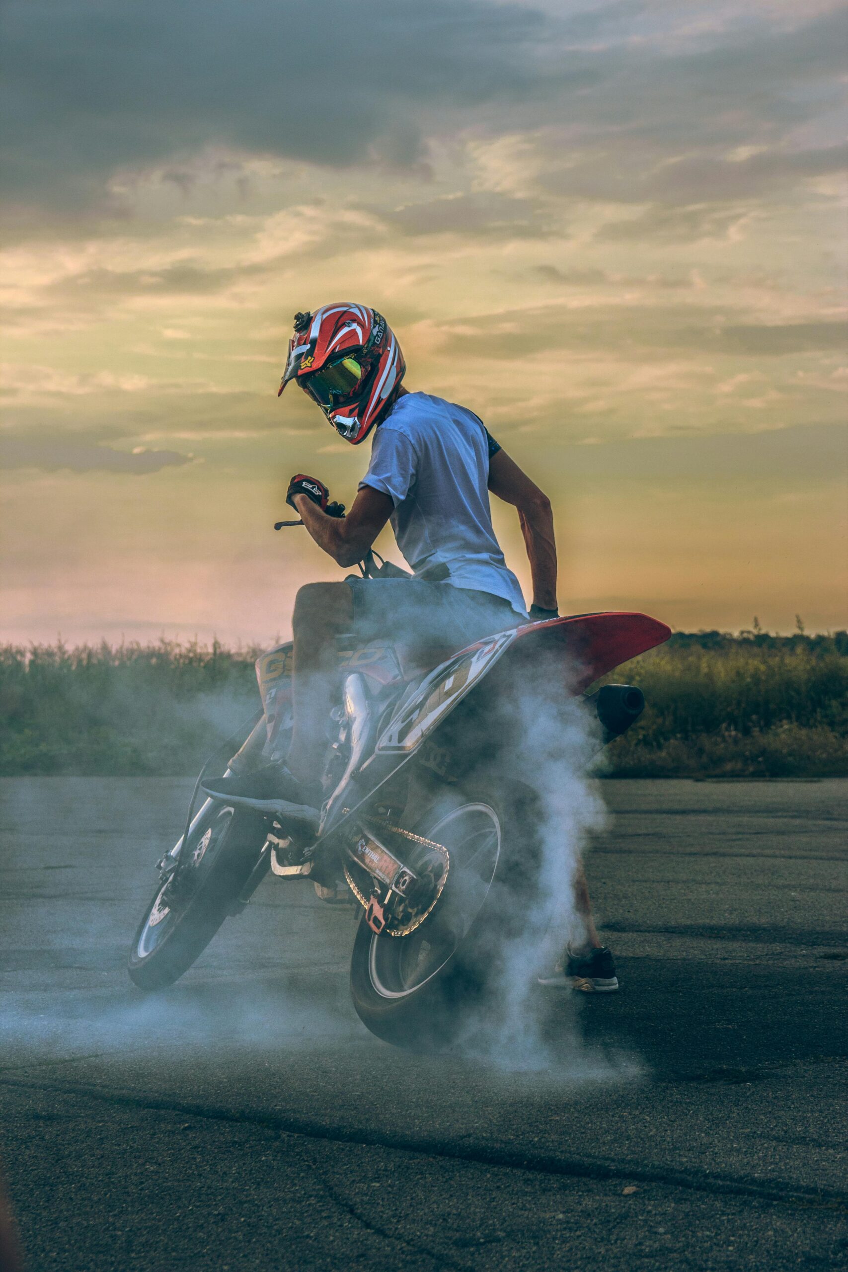 Motorcyclist performing a drift on pavement, creating smoke under a sunset sky.
